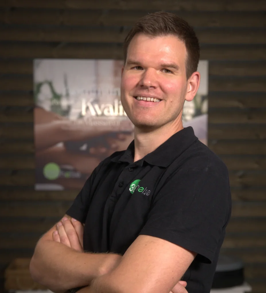 Mann i svart T-skjorte med 360Helse-logo, smilende med armene krysset, innendørs trenings- eller helsemiljø, fokus på helse og velvære, profesjonell helsepersonell, motivasjon og tillit.